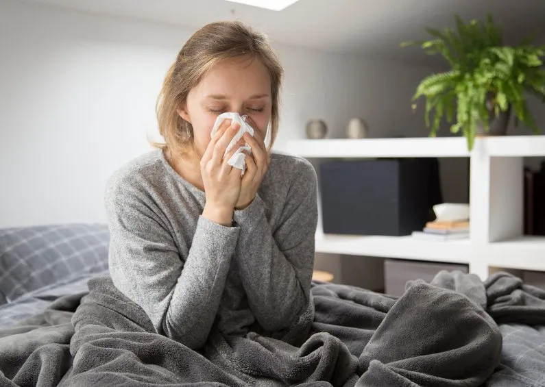 woman with a bad cold is wrapped up in a blanket sitting on her bed blowing her nose